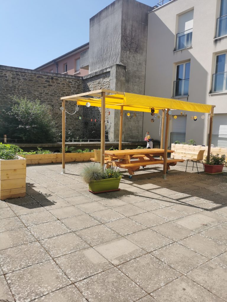 Jardin pédagogique : pergola jaune et tables de pique-nique sur une cour dallée, bacs de plantes en bordure