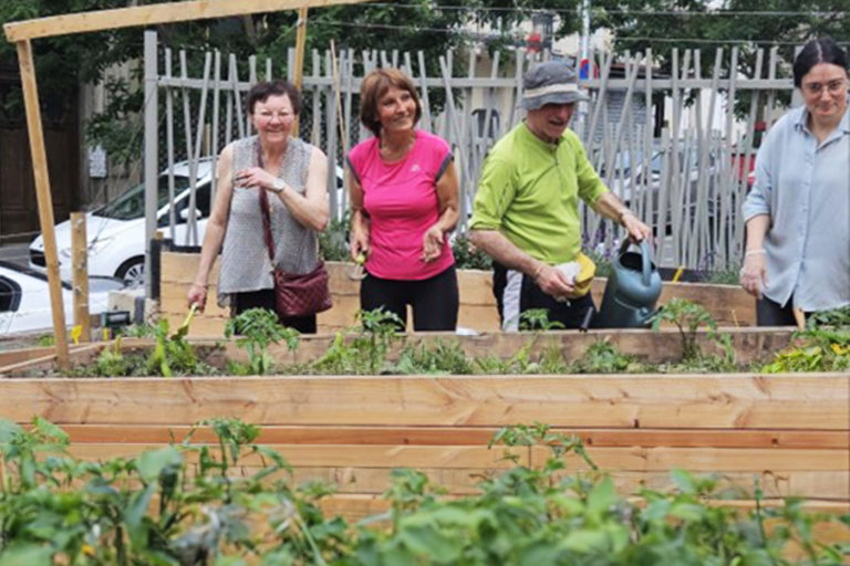 Quatre participants souriants derrière des bacs potagers en bois, arrosoir à la main, lors d'un atelier de jardinage