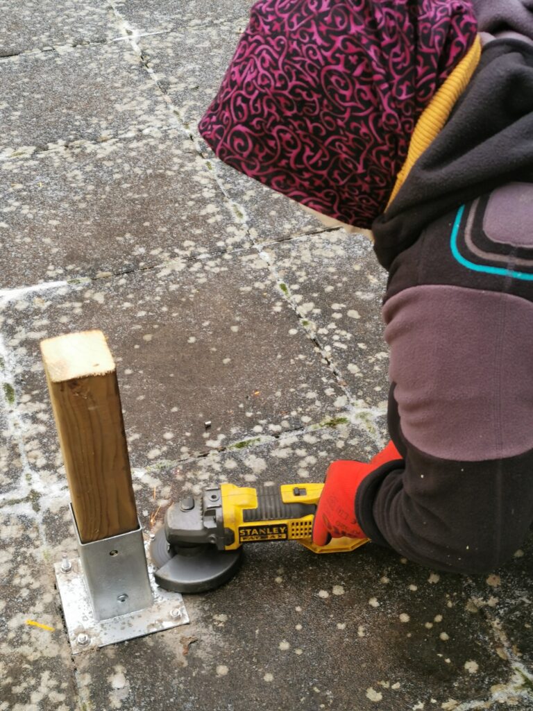 Une personne coiffée d'un foulard à motifs coupe à la meuleuse, dans une gerbe d'étincelles, les tiges d'une platine métallique fixée au sol au pied d'un poteau en bois